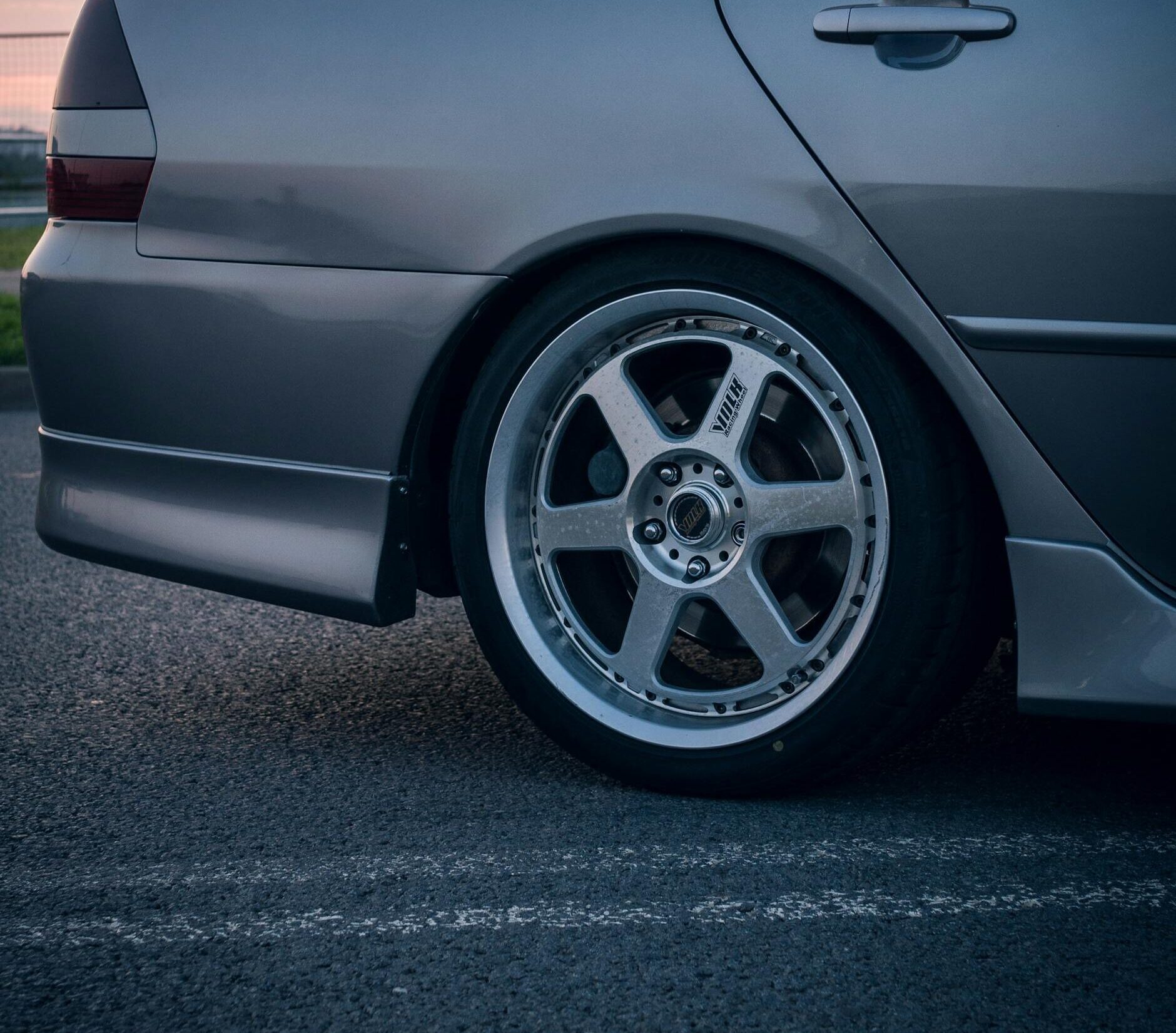 Side view of a silver sedan's alloy wheel parked on the road at twilight.